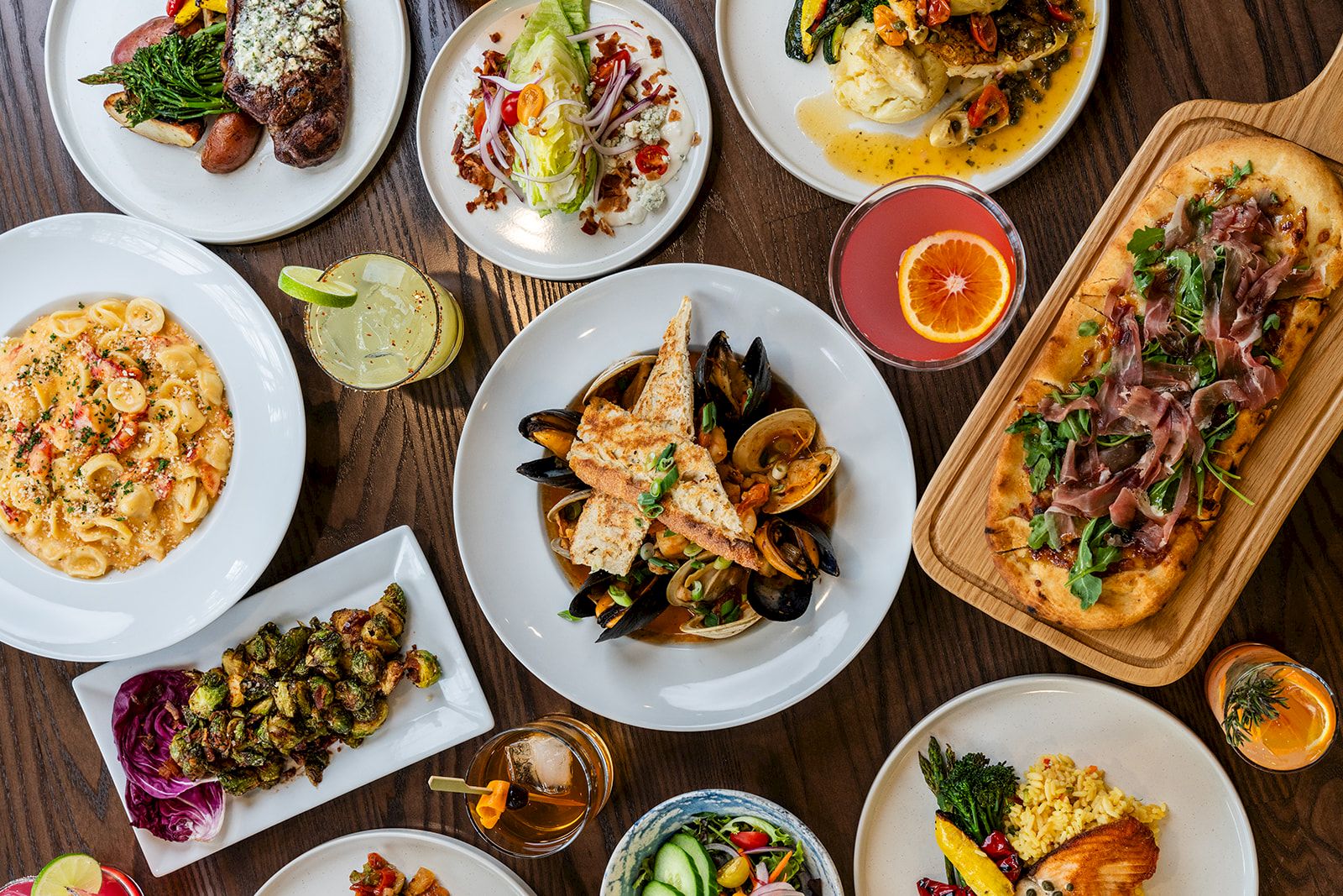 The image depicts a variety of beautifully presented dishes, including seafood, pasta, salad, and flatbread, along with several drinks and garnishments.