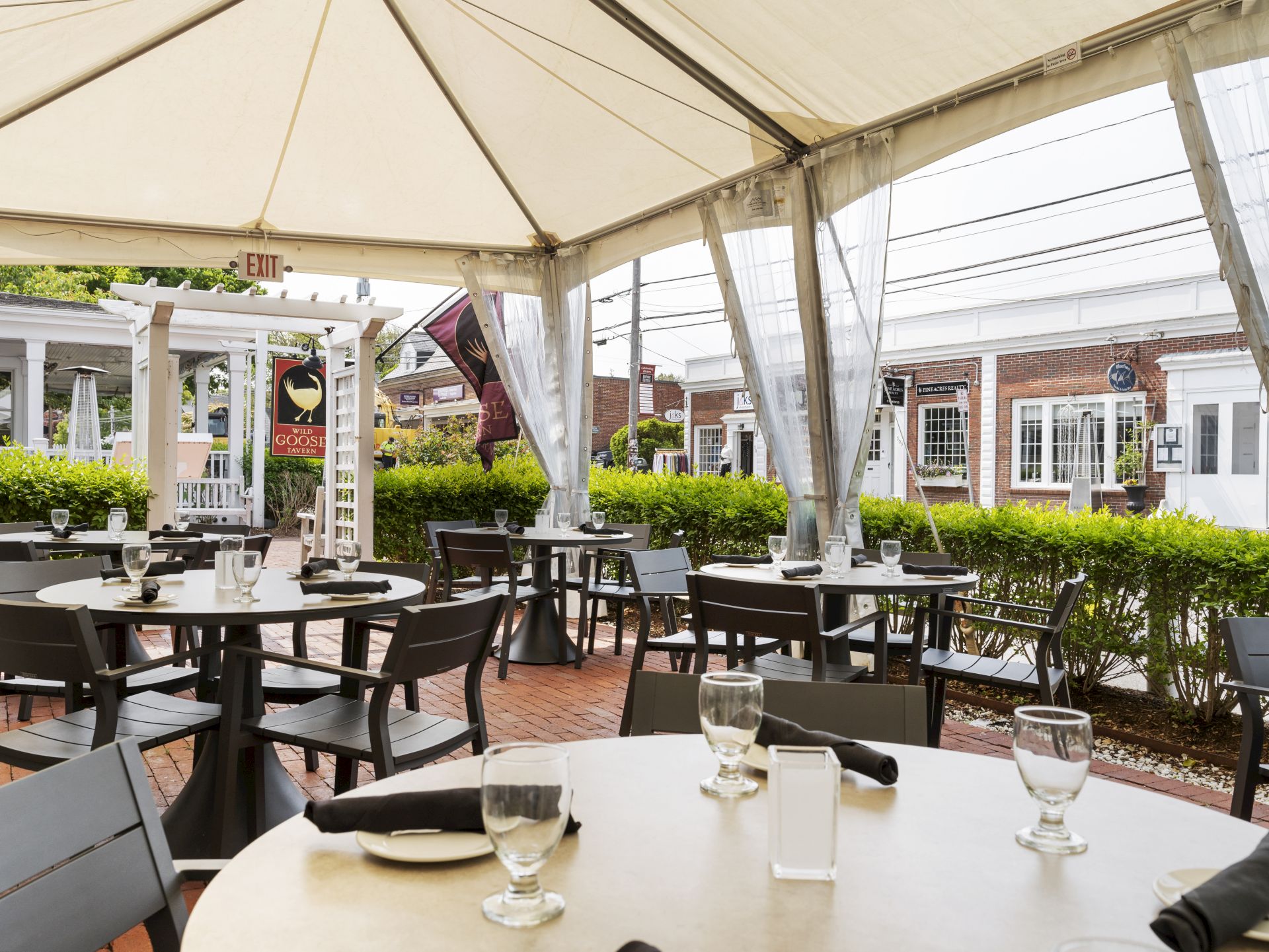 Outdoor dining area under a canopy with set tables, glassware, and black napkins. A trimmed hedge and white structure are visible in the background.