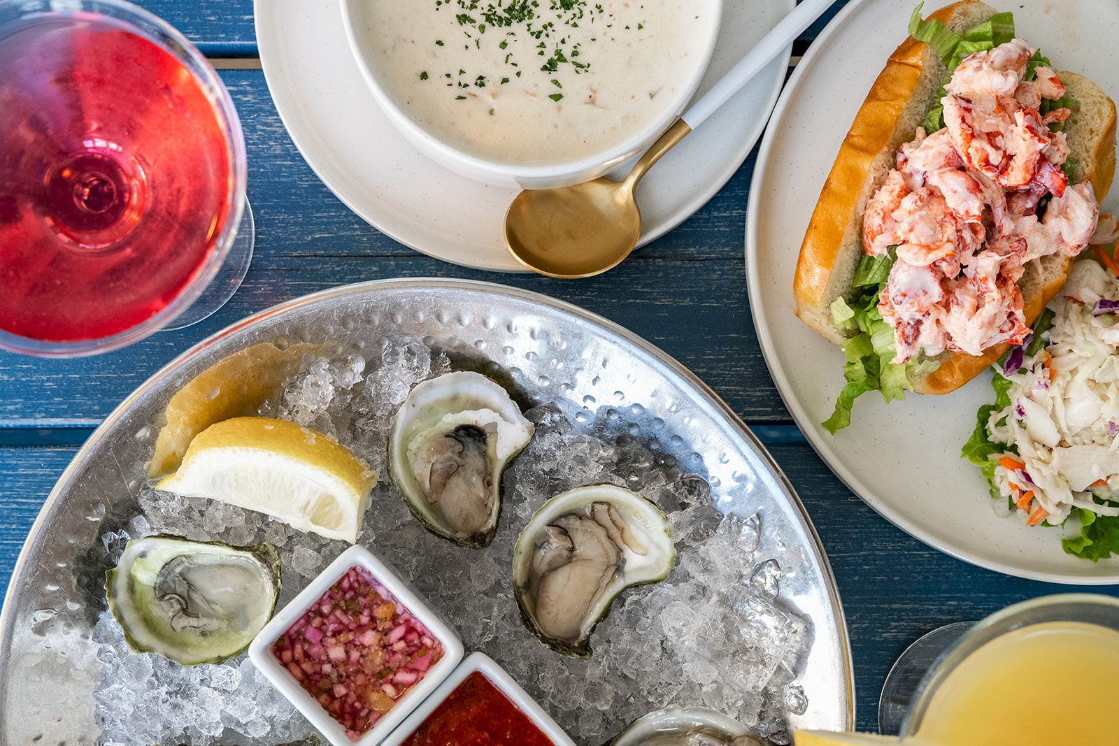 The image shows a seafood meal with oysters on ice, a lobster roll with salad, a bowl of chowder, and glasses of different beverages.