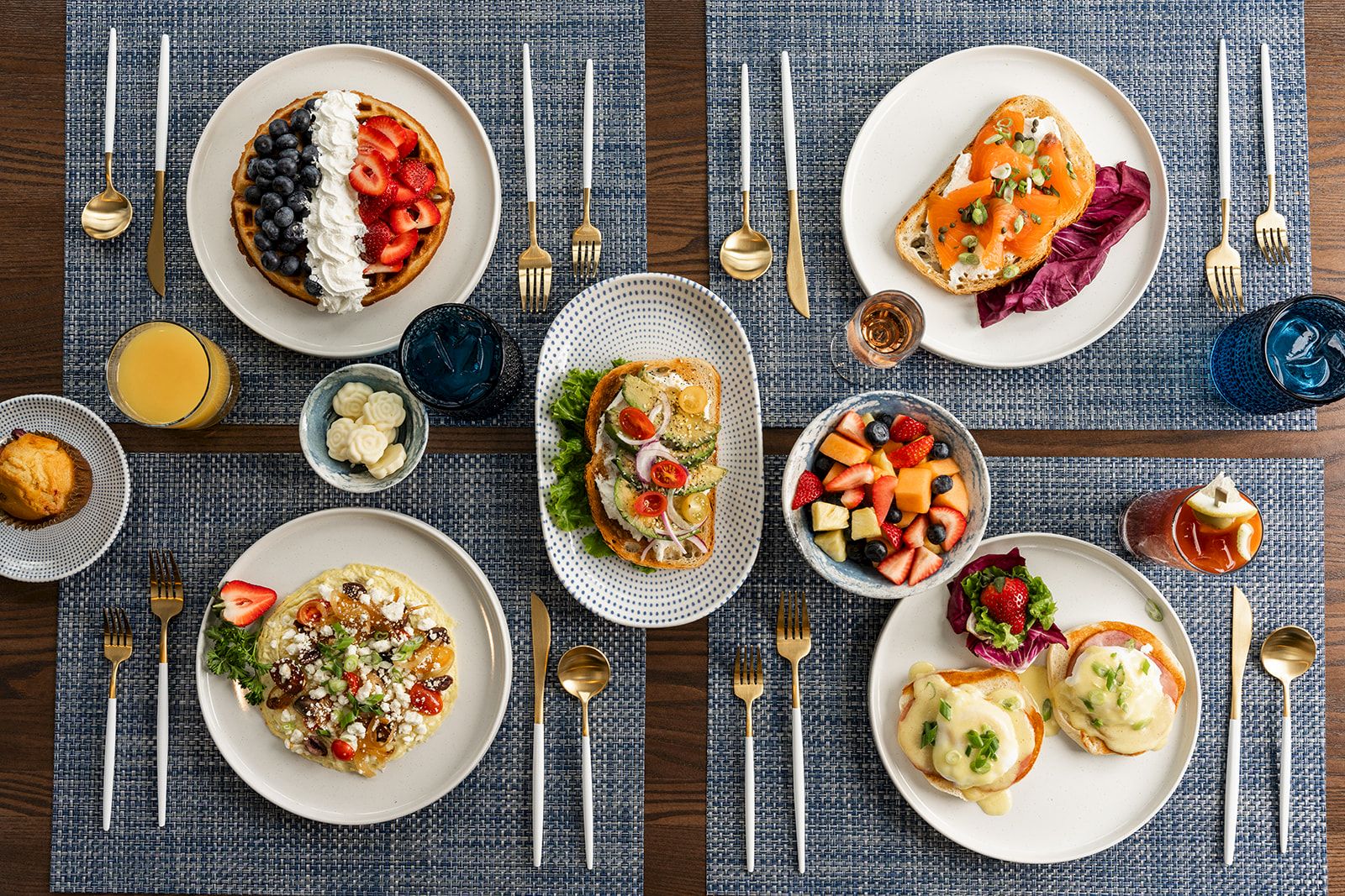 The image shows a table set with various dishes including waffles, salmon toast, fruit salad, and egg dishes, accompanied by drinks and utensils.