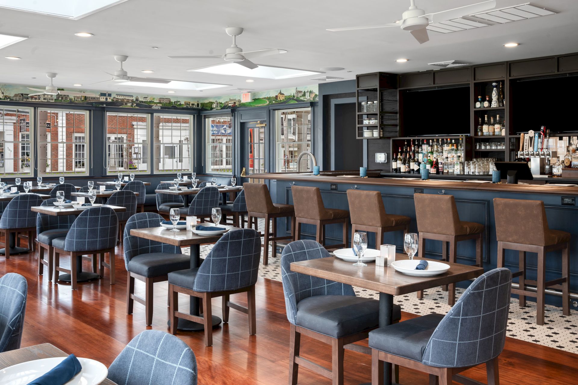 The image shows a modern restaurant interior with neatly arranged tables, blue cushioned chairs, and a fully stocked bar with high stools.