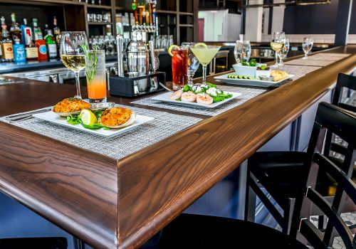 A bar setting with cocktails and a meal displayed on the counter, including shrimp, a drink, and garnished dishes on placemats with stools nearby.
