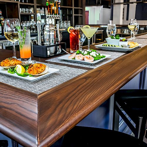A bar setting with cocktails and a meal displayed on the counter, including shrimp, a drink, and garnished dishes on placemats with stools nearby.