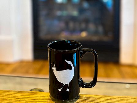 A black mug with a white bird design sits on a wooden table, with a fireplace blurred in the background.