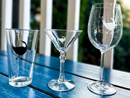 The image shows three different glasses on a blue surface: a pint glass, a martini glass, and a wine glass, each with a bird design.