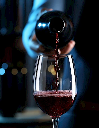 A person pours red wine into a glass, creating a small burgundy splash as the drink fills the stemware, dim bar lighting in the background.