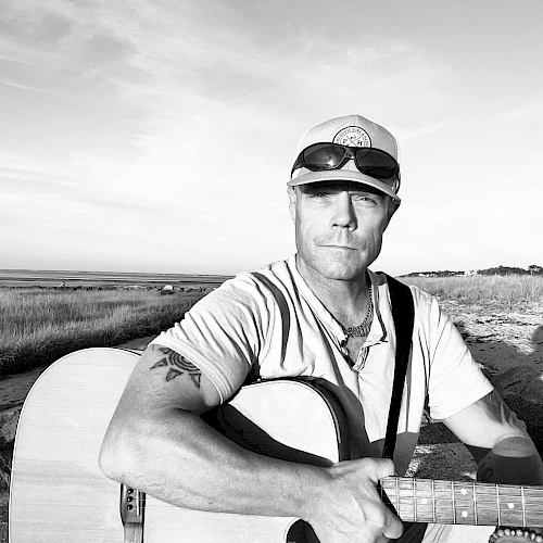 A person wearing sunglasses and a cap is holding a guitar, sitting outdoors by a grassy field under a partly cloudy sky.