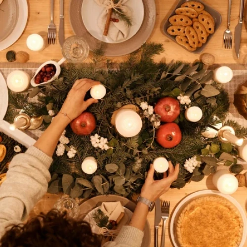 A festive table with a lush centerpiece of greenery and red apples, surrounded by candles, plates, pies, cookies, and seasonal dishes for a warm holiday feast.