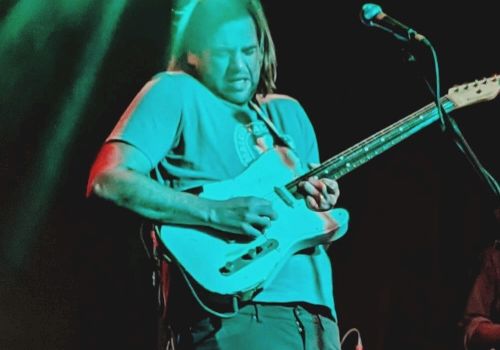 A guitarist wearing a teal shirt plays an electric guitar on stage with a drummer in the background, under colorful lights.