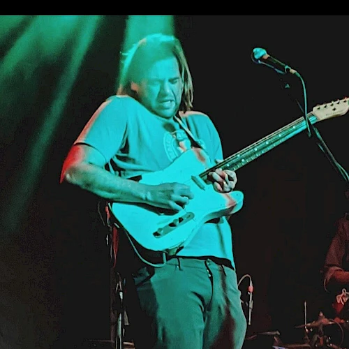 A guitarist wearing a teal shirt plays an electric guitar on stage with a drummer in the background, under colorful lights.