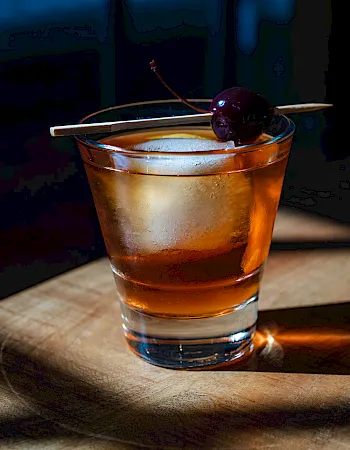 A short glass of amber whiskey on a wooden barrel, with ice and a cherry garnish on a skewer, catching warm light and a shadow.