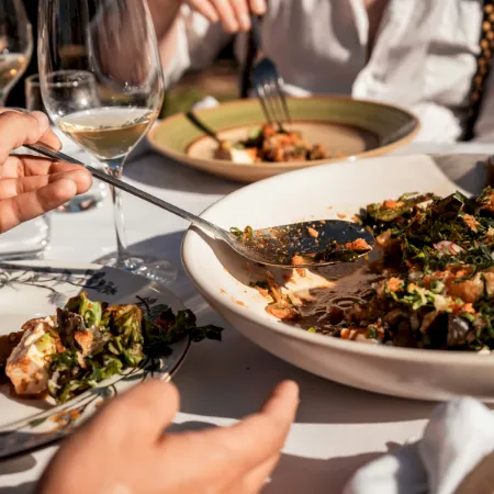 A group dining at a table with wine glasses and bowls of food, as people eat with forks and chop herbs.