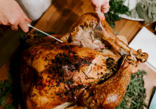 A person carves a roasted, herb-seasoned chicken on a wooden cutting board, slicing into crispy browned skin.