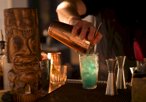 A bartender pours a green cocktail into a glass using a copper shaker, with bar tools and bottles in the background.